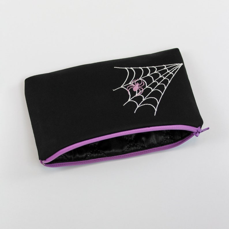 May include: Black zippered pouch with a purple zipper and a white embroidered spider web with a pink spider in the center.