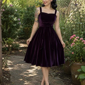 May include: A deep purple velvet midi dress featuring a sweetheart neckline, thick shoulder straps, and a fitted bodice with a matching tie-waist belt. The skirt is full and flares out from the waist, reaching below the knees in a solid dark hue.