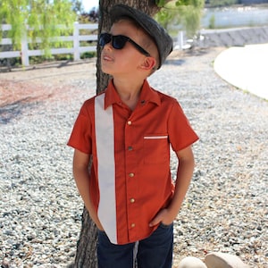 May include: A rust-colored short-sleeve shirt with a vertical white stripe, paired with dark blue jeans. The shirt has a button-down front and a small pocket. The person is wearing sunglasses and a gray cap.
