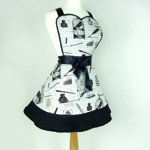 May include: A white apron with a black trim and a black ribbon. The apron has a heart-shaped top and is decorated with a pattern of inkwells, pens, and birds in cages.