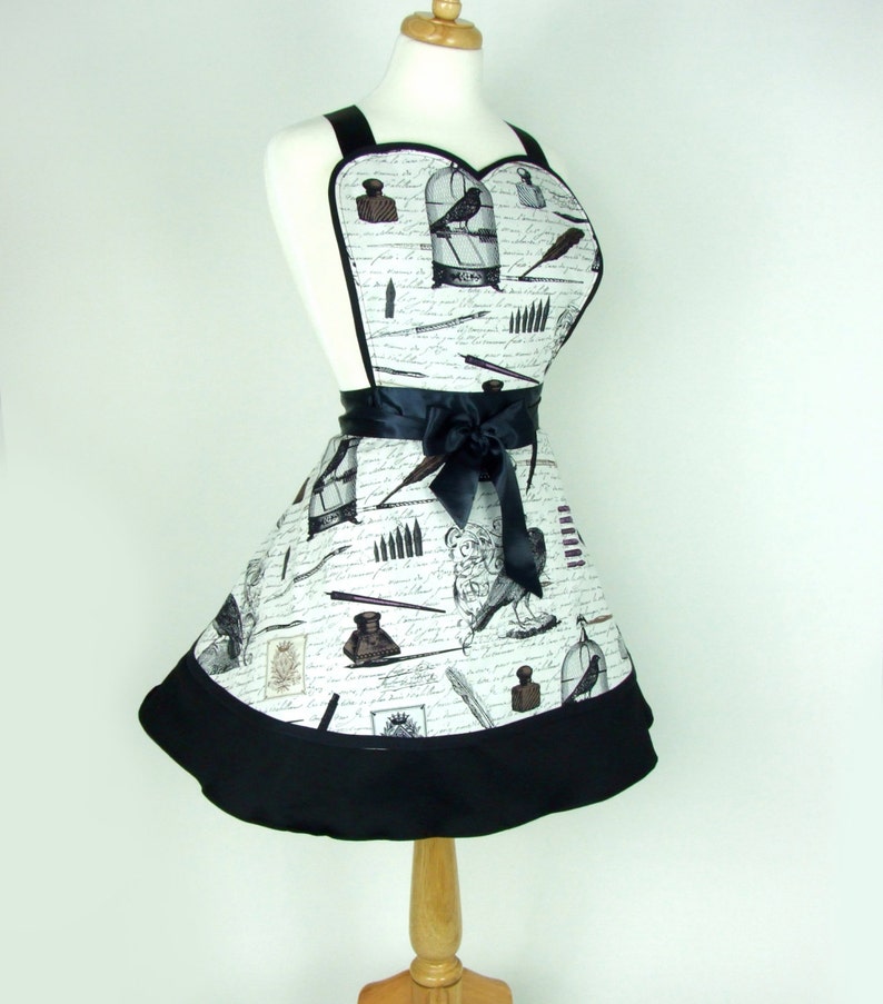 May include: A white apron with a black trim and a black ribbon. The apron has a heart-shaped top and is decorated with a pattern of inkwells, pens, and birds in cages.