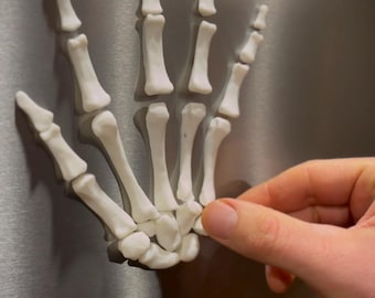 May include: A white, skeletal hand model, likely a decorative item, is affixed to a metallic surface. The hand is detailed, showing individual bones. A person's hand is touching the model, possibly attaching or removing it.