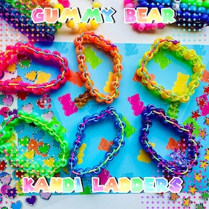 May include: Six colourful beaded bracelets arranged on a blue background featuring gummy bear graphics. The bracelets are constructed with translucent plastic beads in pink, orange, yellow, green, blue, and purple, accented with thin, contrasting elastic cord.