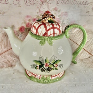 May include: A white ceramic teapot with a red and white plaid lid and a green handle. The teapot is decorated with green leaves and red berries. The teapot is sitting on a white lace tablecloth.