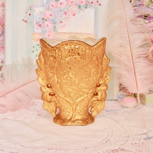 May include: A gold ceramic vase with a decorative floral design. The vase has a wide mouth and a narrow base. It is decorated with a textured, embossed pattern.