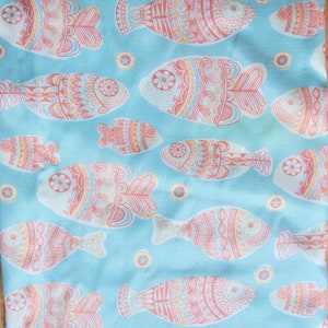 May include: A light blue fabric with a repeating pattern of stylized fish in coral, yellow, and teal. The fish are decorated with intricate patterns and surrounded by small star-shaped accents. The fabric has a textured appearance.