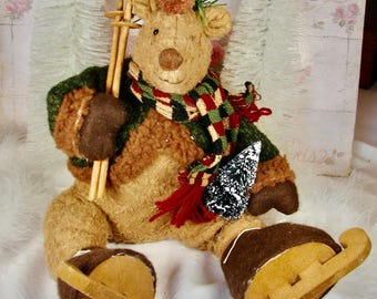 May include: A plush reindeer figurine wearing a green and red scarf, green jacket, and wooden skis. The reindeer has a brown pom-pom hat and is wearing wooden ice skates. A small, decorative Christmas tree is attached to the reindeer.