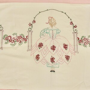 May include: White embroidered pillowcase with a floral design and a woman in a pink dress standing under an archway.