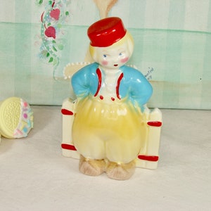 May include: A vintage ceramic figurine of a boy wearing a red hat, blue jacket, and yellow pants. The figurine is standing in front of a white picket fence with red accents. The figurine is hand-painted and has a whimsical charm.