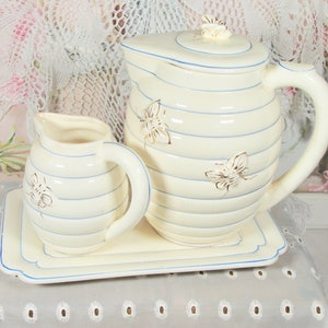 May include: A white ceramic pitcher and creamer set with blue stripes and gold bee accents. The set is on a matching tray with a blue trim.