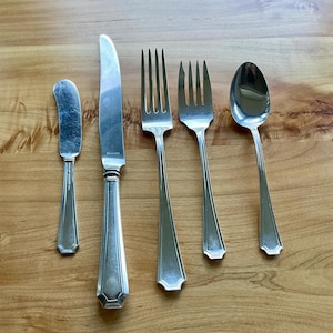 Puede incluir: Un juego de cinco cubiertos de acero inoxidable, que incluye un cuchillo de mantequilla, un cuchillo de mesa, dos tenedores y una cuchara. La cubertería tiene un diseño clásico con un patrón geométrico en los mangos. Las piezas están dispuestas sobre una superficie de madera.