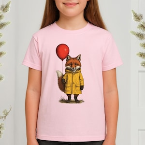 Könnte beinhalten: Hellrosa T-Shirt mit einer Illustration eines Fuchses in einem gelben Regenmantel, der einen roten Ballon hält. Der Fuchs hat einen buschigen Schwanz und steht auf Gras. Das Design ist mittig auf dem T-Shirt platziert.