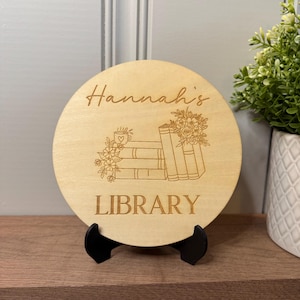 May include: A round wooden sign with the text "Hannah's Library" and a design of stacked books with flowers and a coffee cup.