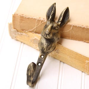 May include: A bronze-colored metal wall hook shaped like a rabbit's head. The hook has a detailed design, with visible ears, eyes, and fur texture. The hook is mounted on a white surface, with a stack of old books in the background.