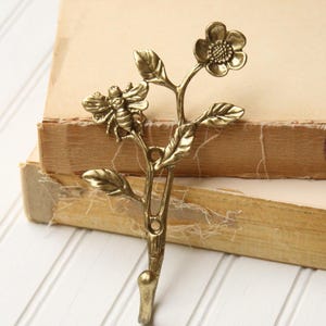 May include: A brass-colored metal wall hook shaped like a branch with a bee and flower. The hook is detailed with leaves and a flower at the top. The hook is resting on top of two old books with yellowed pages.