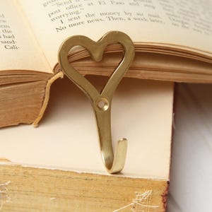 May include: A heart-shaped, brass-colored bookmark is inserted into an open book. The bookmark has a hook at the bottom and a hole near the top. The book's pages are yellowed.
