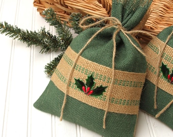 May include: Two green burlap gift bags with a tan stripe and holly berry design, tied with twine. A woven basket and evergreen sprig are in the background. Festive holiday decor.