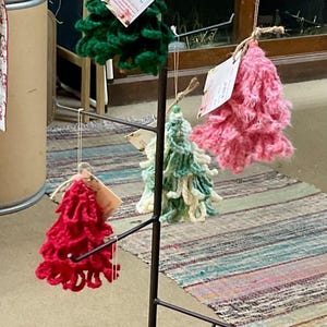 May include: A display of handmade crochet Christmas tree ornaments in various colors, including red, green, pink, and teal. Each tree is suspended from a black metal rack and has a tag attached.
