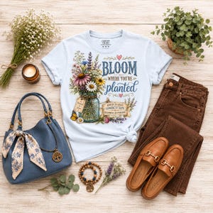 Puede incluir: Una camiseta azul claro con un gr&aacute;fico de un frasco de vidrio lleno de flores silvestres coloridas, una mariposa y el texto "BLOOM WHERE YOU'RE PLANTED" junto a una pancarta que dice "ROOTED IN GRACE GROWING IN FAITH BLOOMING IN PURPOSE."