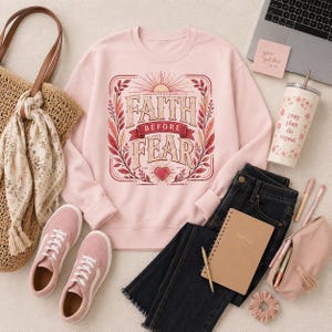 Puede incluir: Una sudadera de cuello redondo rosa claro con un gráfico que incluye el texto "FAITH BEFORE FEAR" en una fuente decorativa, rodeado de hojas estilizadas, un sol naciente y un corazón en tonos rojos, naranjas y crema.