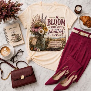 Puede incluir: Una camiseta de color crema con un gr&aacute;fico de un frasco de vidrio lleno de flores silvestres y el texto "BLOOM WHERE YOU'RE planted" junto con "ROOTED IN GRACE GROWING IN FAITH BLOOMING IN PURPOSE" impreso en una fuente granate.