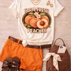Puede incluir: Una camiseta color crema con un gráfico de melocotones y el texto FARM FRESH PEACHES, combinada con pantalones cortos naranjas, un cinturón marrón, sandalias marrones, una mochila marrón con un lazo blanco, un collar dorado y pendientes dorados.