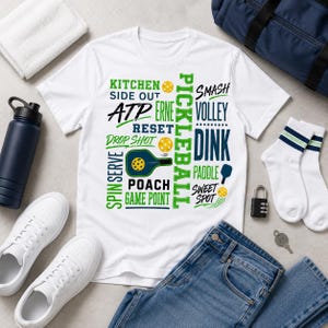 Puede incluir: Una camiseta blanca de manga corta con un estampado gráfico de terminología de pickleball que incluye las palabras Kitchen, ATP, Erne, Reset, Drop Shot, Spin Serve, Poach, Game Point, Smash, Volley, Dink, Paddle, Sweet Spot y Pickleball.