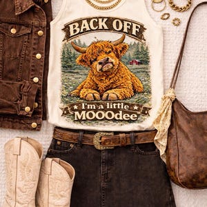Puede incluir: Una camiseta sin mangas color crema con un gráfico de una vaca Highland y el texto BACK OFF e I'm a little MOOOdee, junto con una chaqueta vaquera marrón, una falda vaquera negra, un cinturón de cuero marrón, un bolso y botas vaqueras.