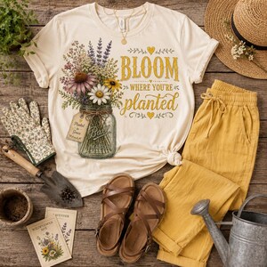 Puede incluir: Una camiseta de color crema con un gr&aacute;fico de un frasco de vidrio lleno de flores silvestres coloridas y el texto "BLOOM WHERE YOU'RE planted" en letras amarillas y marrones. El dise&ntilde;o incluye una peque&ntilde;a etiqueta y corazones decorativos.