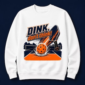 Puede incluir: Una sudadera blanca de cuello redondo con un gráfico de una pista de pickleball, una pala y una pelota naranja con agujeros negros. El texto "DINK, don't think!" está impreso en letras negritas de color azul marino y naranja en el frente.