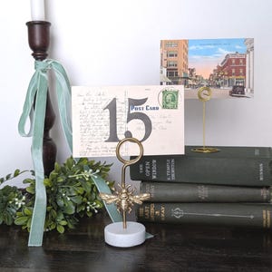 May include: A decorative display featuring a vintage postcard held by a gold-colored bee-shaped holder on a marble base. A second postcard is held by a simple gold wire holder. Green books and a candle holder with a green ribbon are also present.