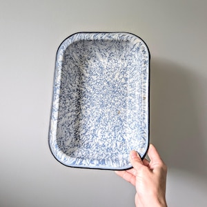 May include: Rectangular enamelware baking dish with a blue and white speckled pattern. The dish has a black rim and is held against a gray background. This vintage-style dish is suitable for baking and serving.