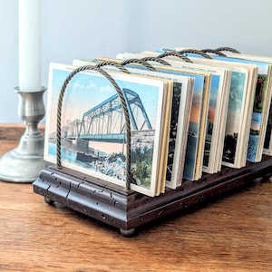 May include: A vintage postcard holder made of dark wood with a metal wire frame. The holder is filled with postcards featuring images of bridges and scenic landscapes.