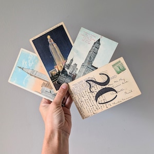 May include: A hand holds a collection of vintage postcards featuring architectural landmarks. The postcards showcase images of tall buildings, with one bearing a large stylized number and handwritten text. The color palette includes blues, greens, and beige.