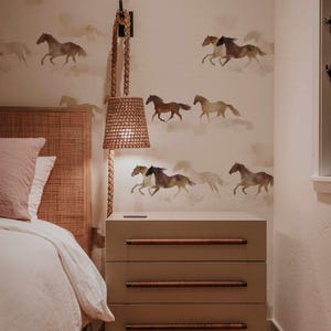 May include: Bedroom interior with a beige nightstand featuring three drawers with wooden handles. The wall is adorned with a horse-themed wallpaper. A woven lamp hangs from the wall, and a bed with a rattan headboard is visible.