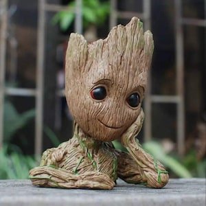 Pode incluir: Um vaso de cerâmica marrom e verde em forma do personagem Groot do filme Guardiões da Galáxia. Groot está sentado com o queixo apoiado na mão e tem um pequeno sorriso no rosto. O vaso tem um furo no topo para plantar.