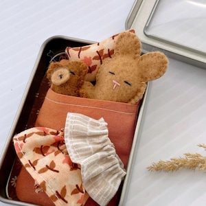 May include: A small brown bunny and bear are sleeping in a bed made of fabric. The bed is in a metal tin with a floral pattern.