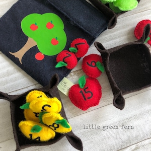 May include: A felt apple tree with a felt bag and four felt baskets filled with felt apples and pears with numbers on them. The apples are red and the pears are yellow. The numbers on the apples and pears range from 1 to 15.