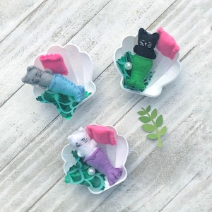 May include: Three felt cat mermaid toys in white, gray, and black, each with a pink pillow and a green, blue, or purple tail. The toys are in white seashell-shaped containers with green and white patterned fabric inside.