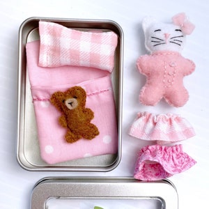 May include: A silver tin with a pink and white gingham pillow, a pink blanket with white polka dots, a brown teddy bear, a white cat with pink pajamas and a pink gingham skirt, and a pink skirt with a floral pattern. The tin also has a clear window with a green fern and the text "little green fern".