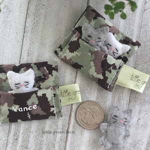 May include: Two small felt cat toys are sleeping in a camouflage fabric pouch with the name Vance embroidered on it. The pouch is next to a larger camouflage pouch with two felt cats sleeping inside. The larger pouch has a green tag with the text "little green fern" on it.