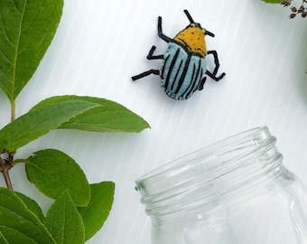 May include: A handmade felt beetle with a yellow head, blue and black striped body, and black legs. The beetle is placed on a white surface with green leaves and a clear glass jar.