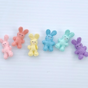 May include: Six felt bunny rabbit figurines in pastel colors: pink, light pink, yellow, light blue, blue, and purple. The bunnies are all facing the right side of the image.
