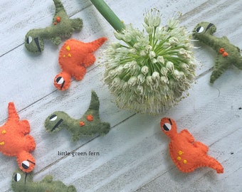 May include: A collection of felt dinosaur toys in green and orange colors. The dinosaurs are arranged on a white wooden surface with a green onion flower in the background. The text "little green fern" is visible in the image.
