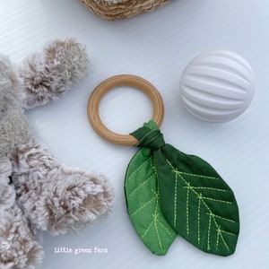 May include: A wooden teething ring with a green fabric leaf attached. The leaf has a light green and dark green color gradient and is stitched with light green thread.