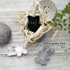 May include: Three felt cat figurines, one black cat inside a speckled gray egg shell, one white cat and one gray cat lying on a white wood surface. The text "little green fern" is visible below the egg.