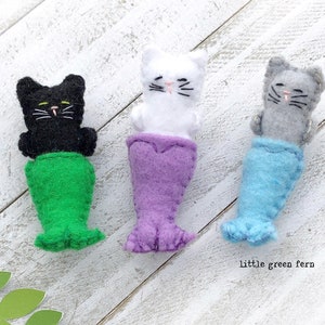 May include: Three felt cat mermaid toys. The first cat is black with a green tail, the second is white with a purple tail, and the third is gray with a blue tail.  The text "little green fern" is at the bottom of the image.