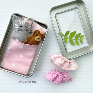 May include: A small, open metal tin contains a gray felt cat with a pink bow, a brown teddy bear, and a pink polka-dot blanket. Two miniature pink skirts and a green leaf are also present. The text "little green fern" is visible.