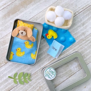 May include: A miniature felt dog in a tin bed with a blue blanket featuring yellow ducklings. Beside it are a basket of white balls, a yellow duck, and a tiny book labeled "Stories". A green leaf and a sticker are also present.