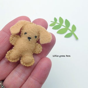 May include: A small, brown felt dog with black eyes and a black nose. The dog is sitting in a hand, and there is a green paper fern in the background. The text "little green fern" is written below the fern.